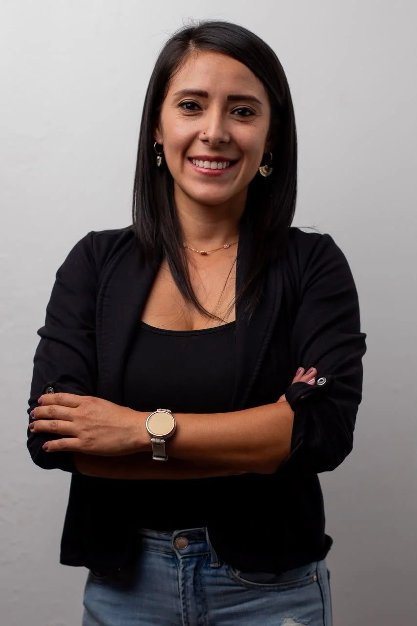 A woman with dark hair, smiling, wearing a black blazer, a black top, jeans, a watch, and earrings, standing against a plain light grey background.