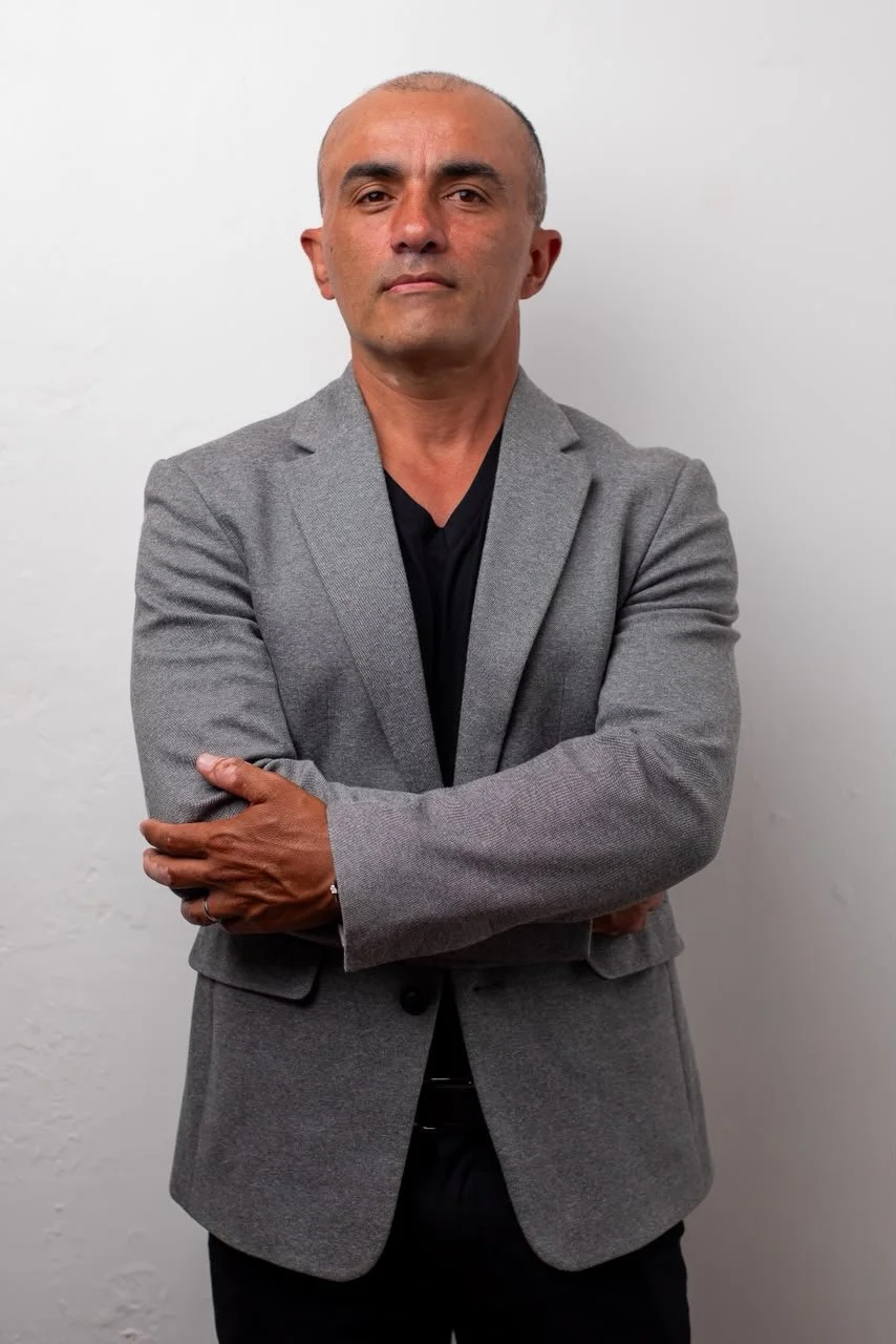 Man with a serious expression, wearing a gray blazer and black shirt, standing with arms crossed against a plain white wall.
