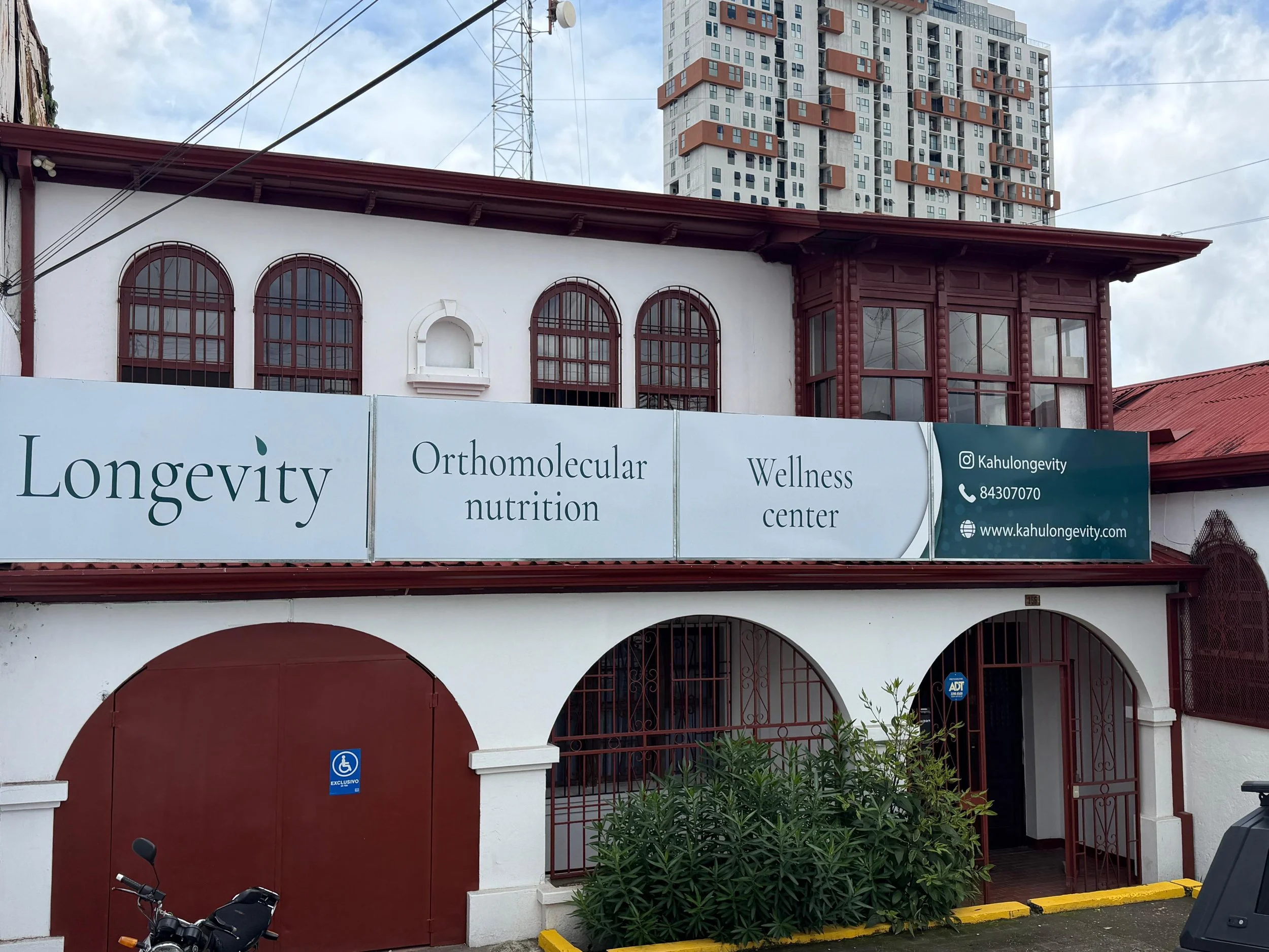 The image shows the front of a building with a sign that reads 'Longevity Orthomolecular Nutrition Wellness Center'. The building has arched windows with brown frames and a red roof. There is a blue handicap parking sign on the gate. In the background, there is a tall modern high-rise building with numerous windows and balconies. The sky is partly cloudy.