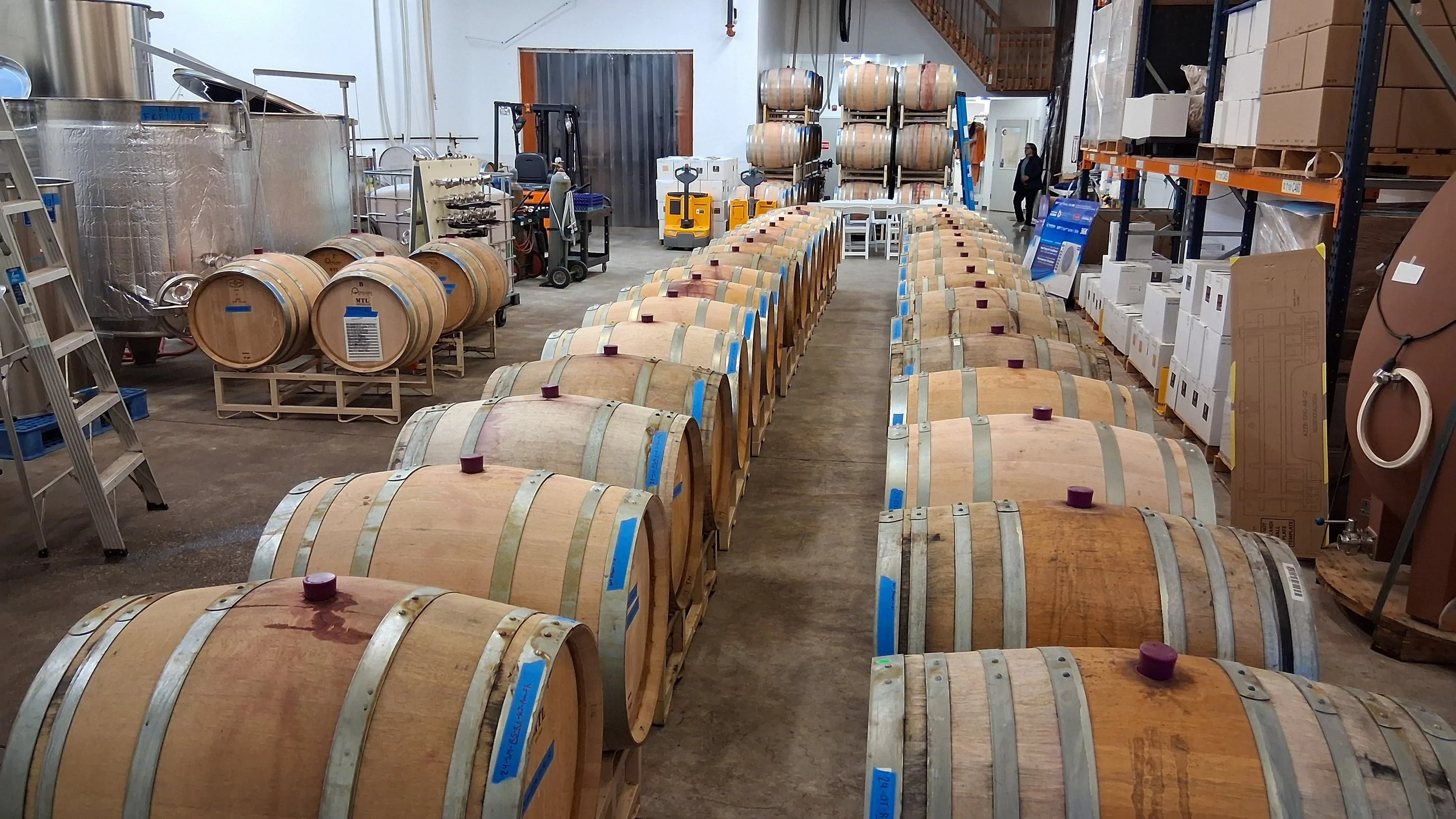 Rows of wooden wine barrels lined up in a warehouse with shelves, storage tanks, and equipment visible in the background.