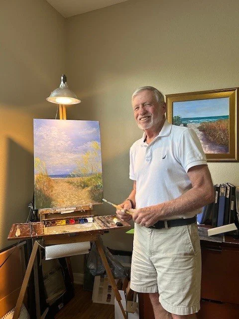 Bob Vogel standing in a home art studio, smiling, holding a paintbrush, next to an easel with a landscape painting of a sunny field, with a lamp, books, and another landscape painting on the wall.