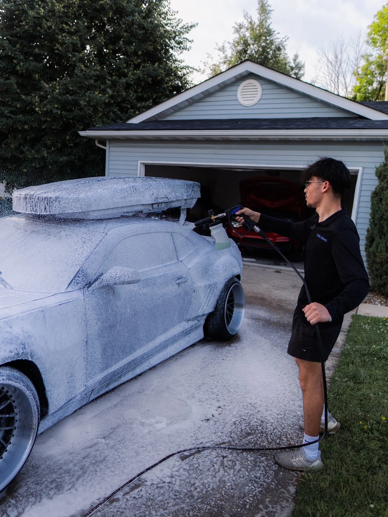A young man wearing black shorts, a black jacket, glasses, and white sneakers is washing a silver car with soap and water using a pressure washer in a driveway next to a house with an open garage. The house has a light blue exterior, and there are tr