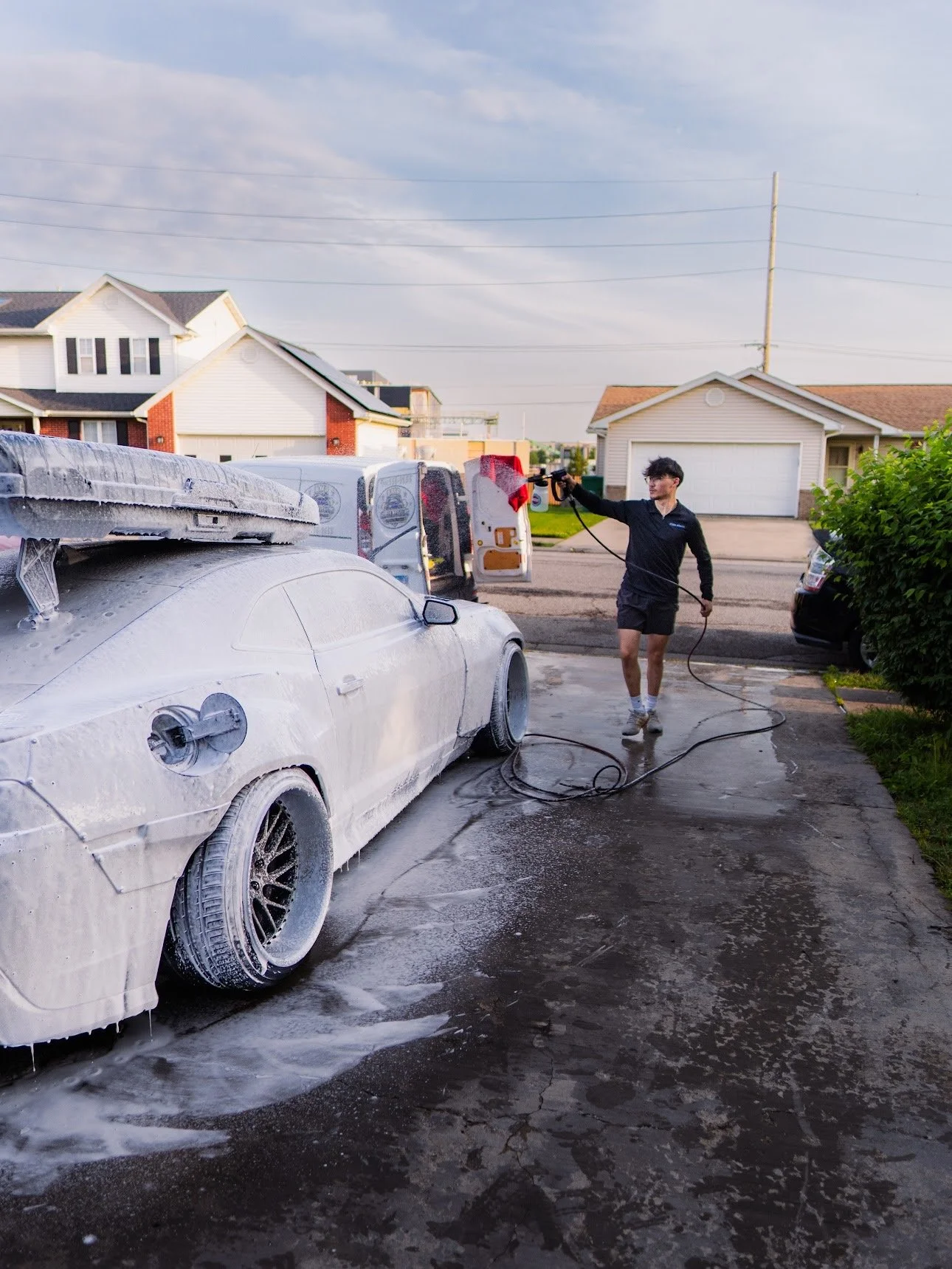 Man washing a white sports car covered in soap suds and foam outside in a driveway with houses and a cloudy sky in the background.