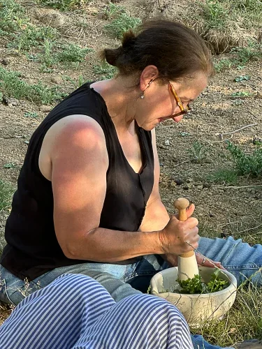 A woman with glasses and a black sleeveless top is sitting outdoors, using a pestle and mortar to crush herbs, with a bowl of herbs nearby.