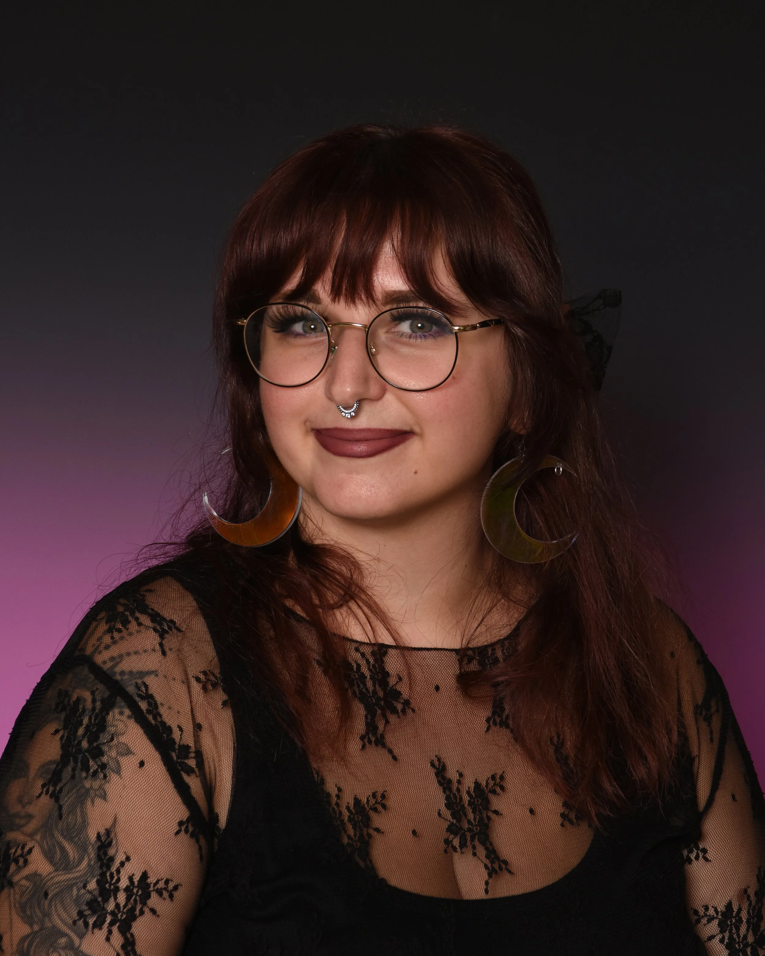 A woman with reddish-brown hair, wearing round glasses, a septum piercing, large crescent-shaped earrings, and a black lace top, smiles at the camera against a dark purple background.