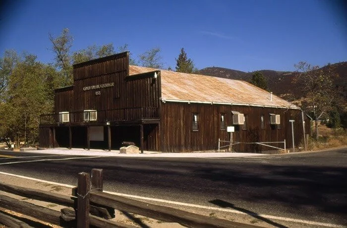 Old wooden building with a sign reading 'COWBOY & COWGIRL HALL' under a clear blue sky, surrounded by trees and distant mountains.