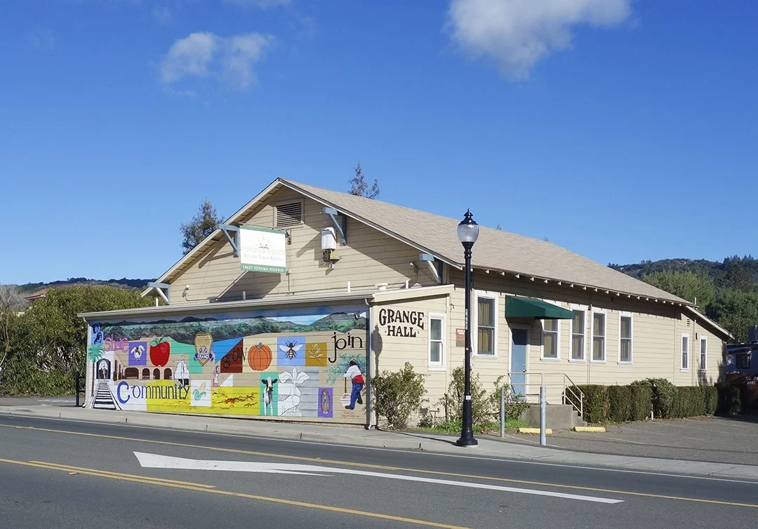 A beige community center building with a colorful mural on the front, a black lamppost, and a blue door. The mural includes various images and the word "community". The building has a sign reading "Grange Hall" and a green awning over a window, with trees and a blue sky in the background.