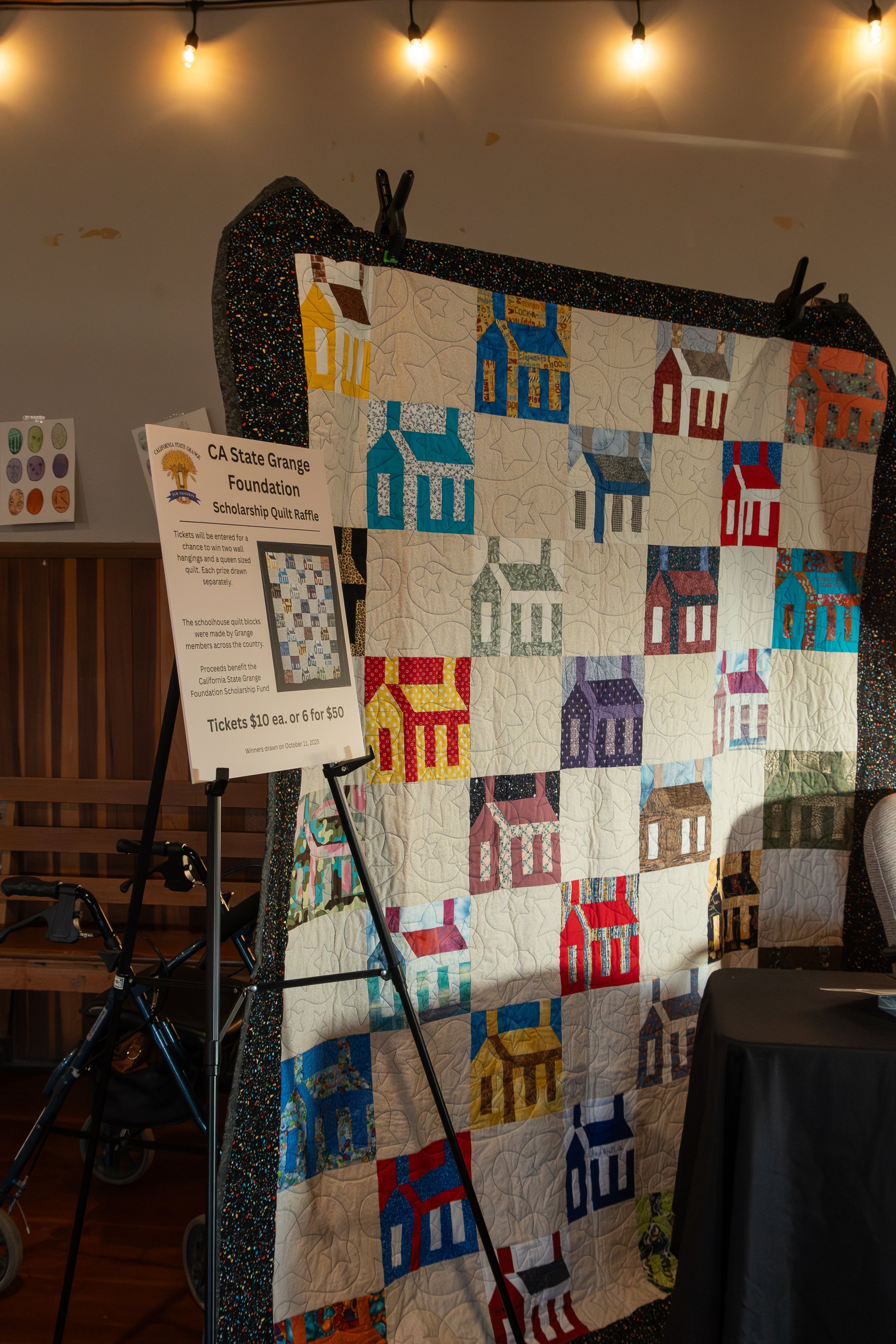 A quilt featuring colorful houses with various patterns, hanging on a display at a fundraising event, with a sign about a raffle and string lights above.