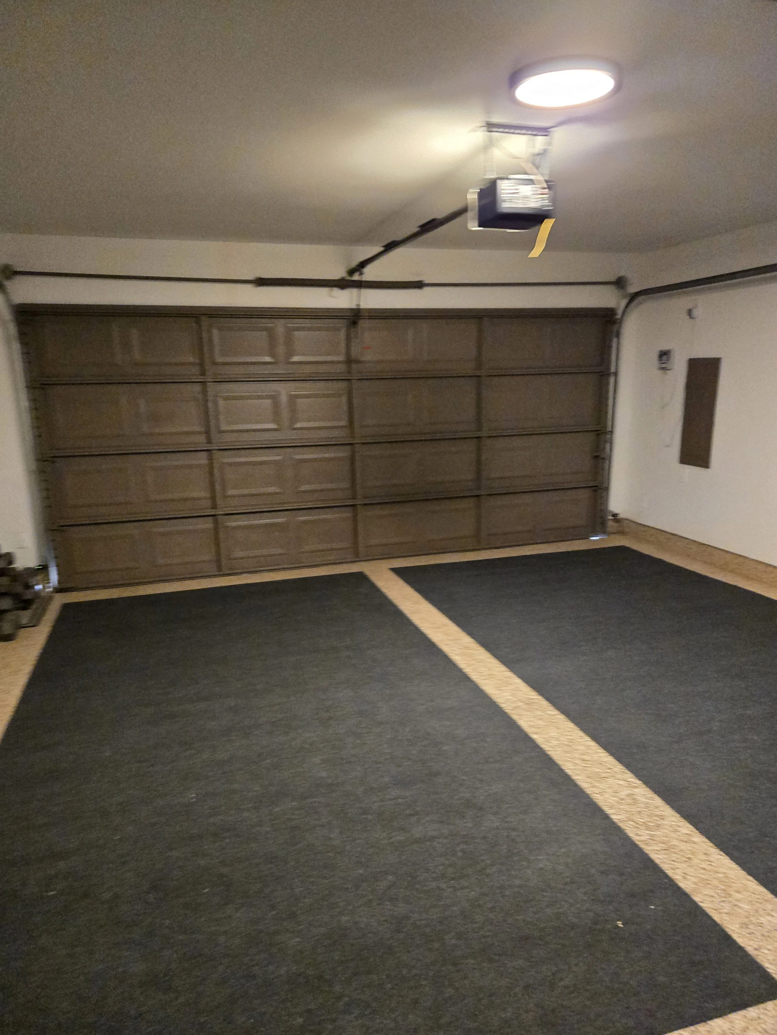 Empty garage with two parking spaces, dark rubber mats, a closed brown garage door, and a ceiling light.
