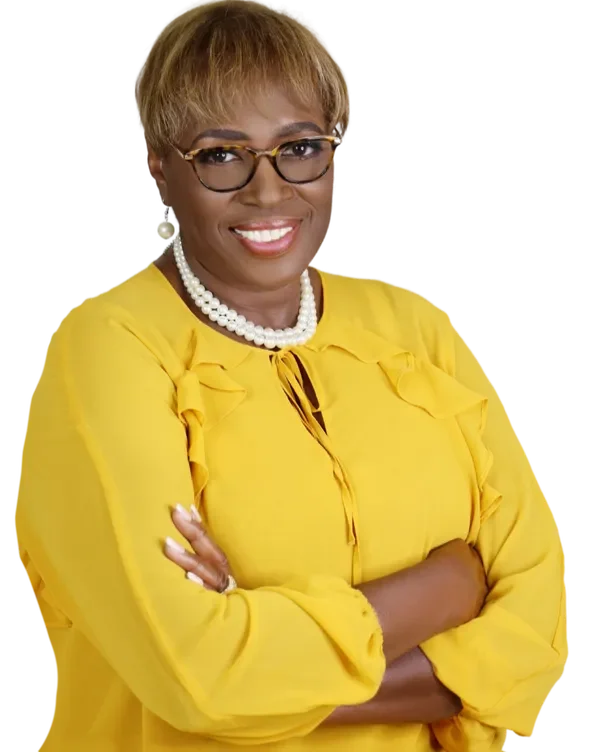 An African American woman with short light brown hair and glasses, wearing a yellow blouse with ruffle details and a pearl necklace, smiling with her arms crossed.