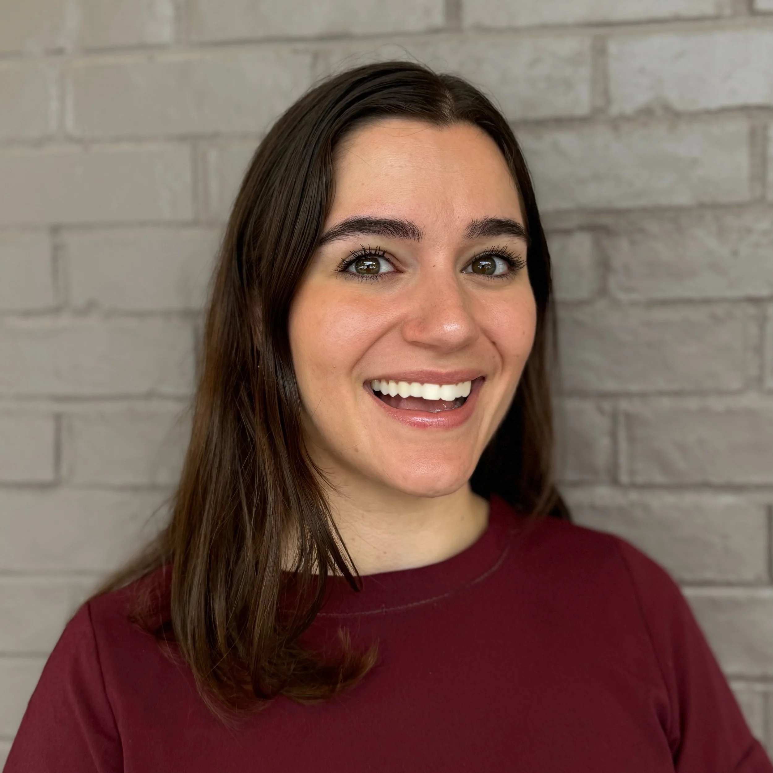 A woman with long brown hair, smiling with white teeth, wearing a burgundy shirt, standing in front of a light gray brick wall.
