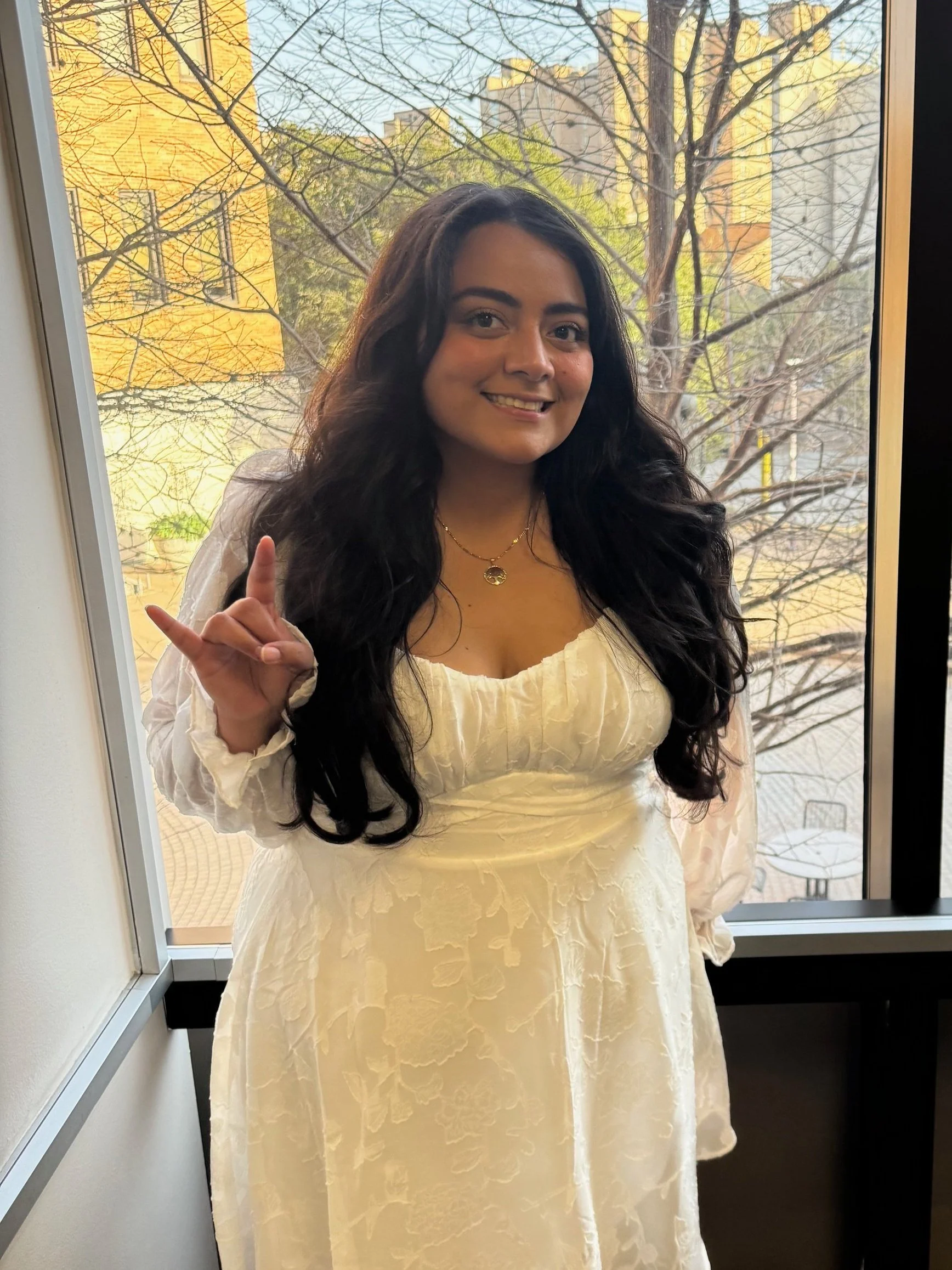 A young woman with long dark wavy hair smiling and holding up her hand forming a sign with her index finger and pinky finger extended, standing near a window with a view of trees and buildings outside.