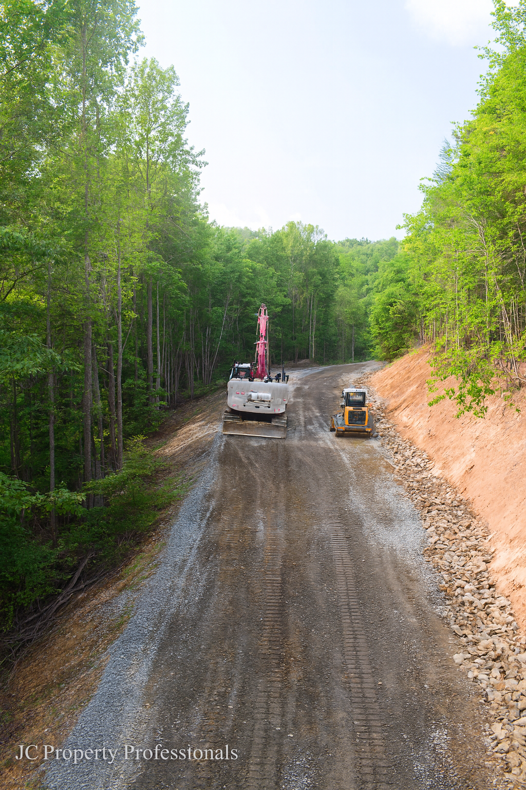 Construction workers leveling a dirt road in a forest area with green trees on both sides.