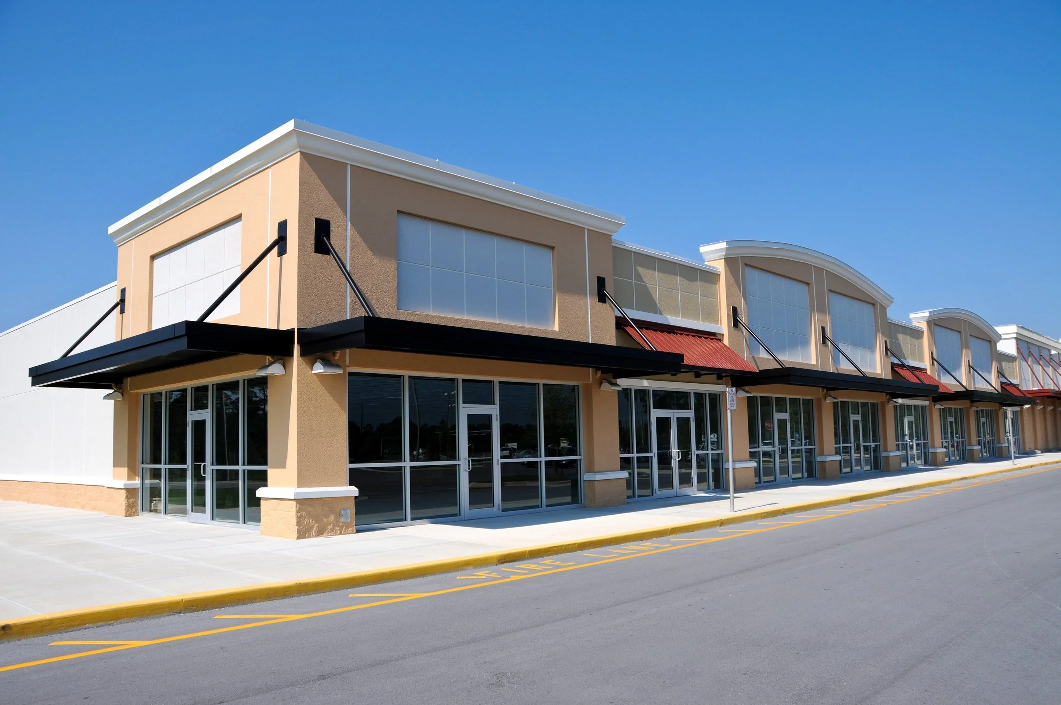 Empty storefront shopping plaza with large windows, awnings, and parking spaces, sunny day with clear blue sky.