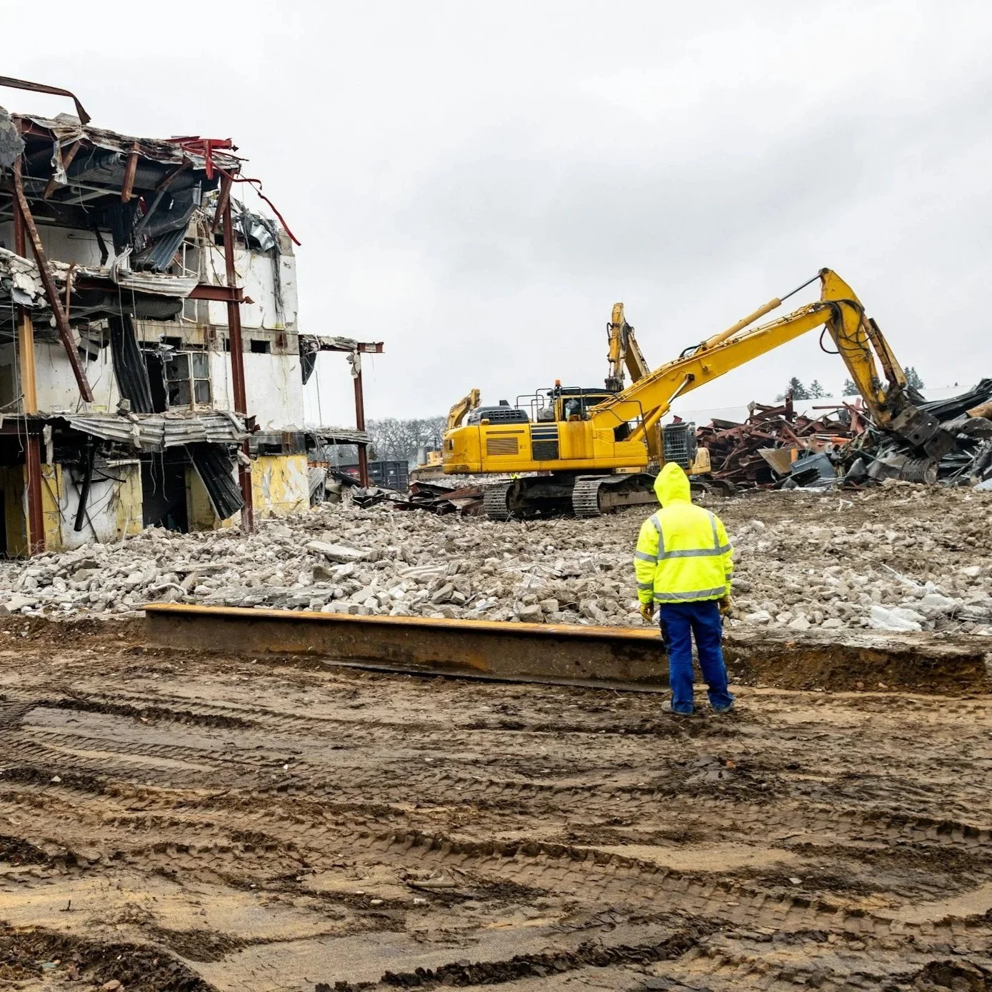 A large yellow excavator demolishing a multi-story concrete building with debris and dust around.