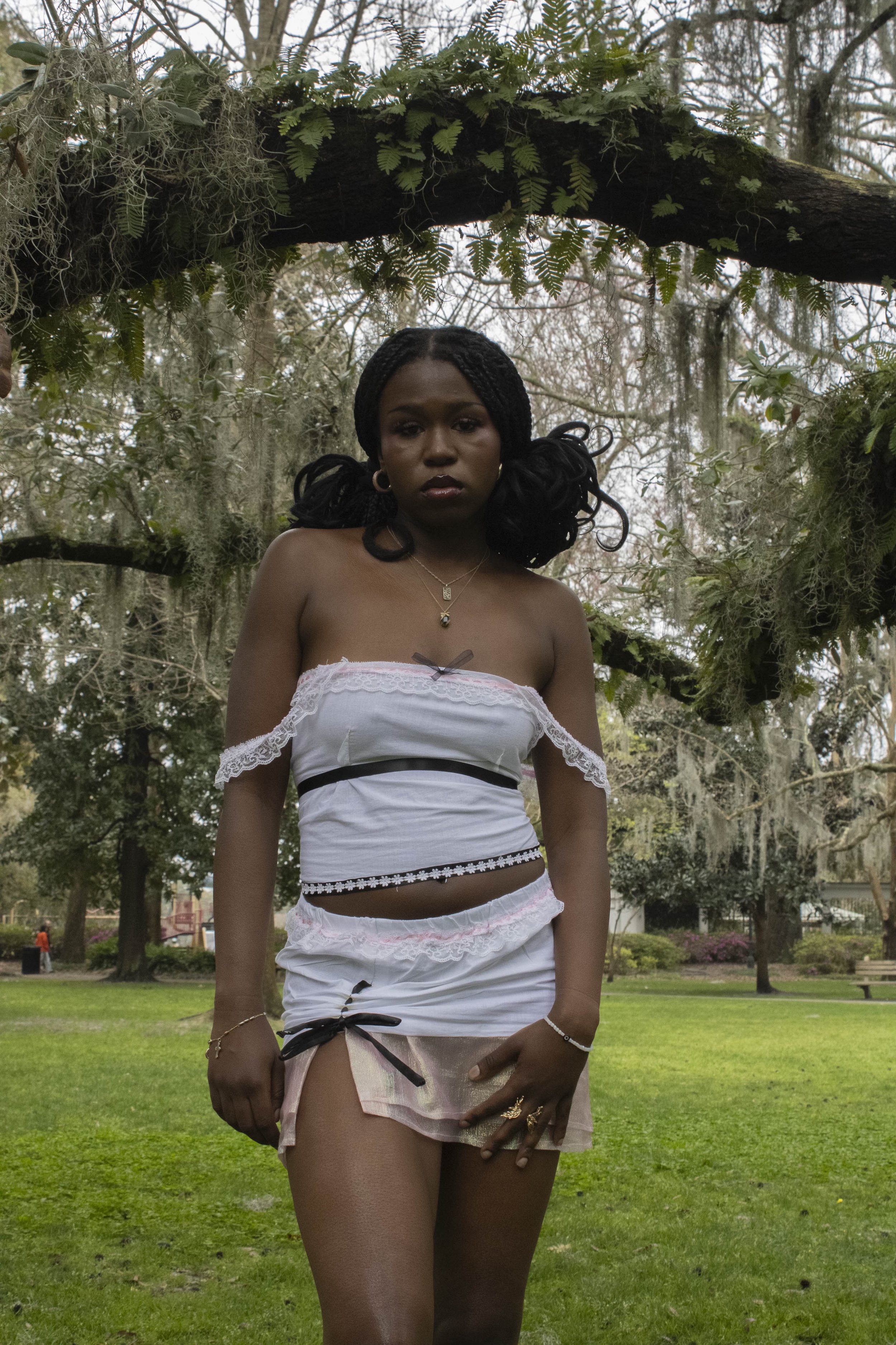 A woman standing outdoors in a park, underneath a tree with moss and ferns, wearing a white off-shoulder top and a short skirt, with her hand resting on her thigh, looking directly at the camera.
