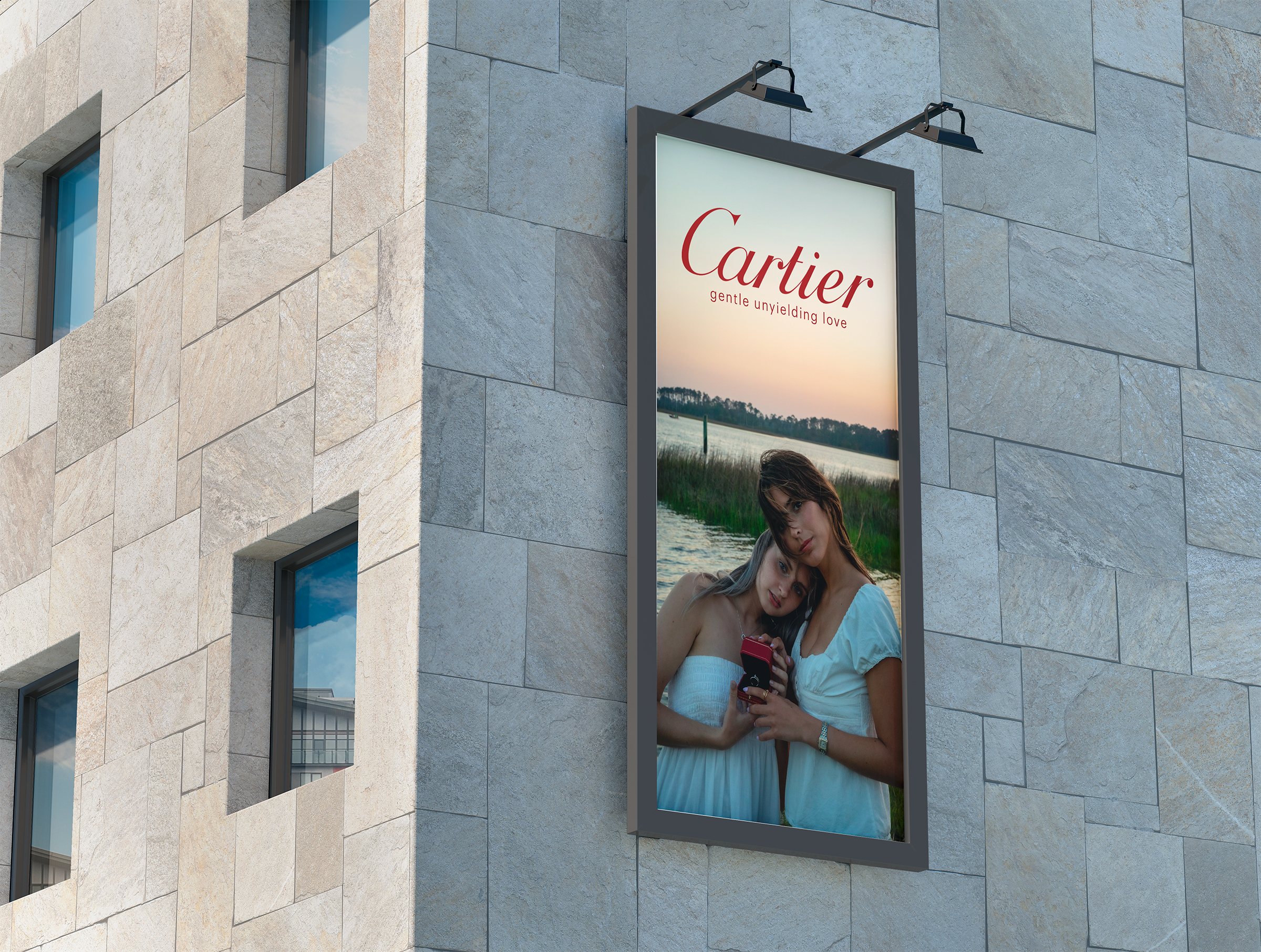 Vertical billboard on a stone building featuring an advertisement for Cartier; the ad depicts two women in white dresses by a lake at sunset, with one woman holding a red box.