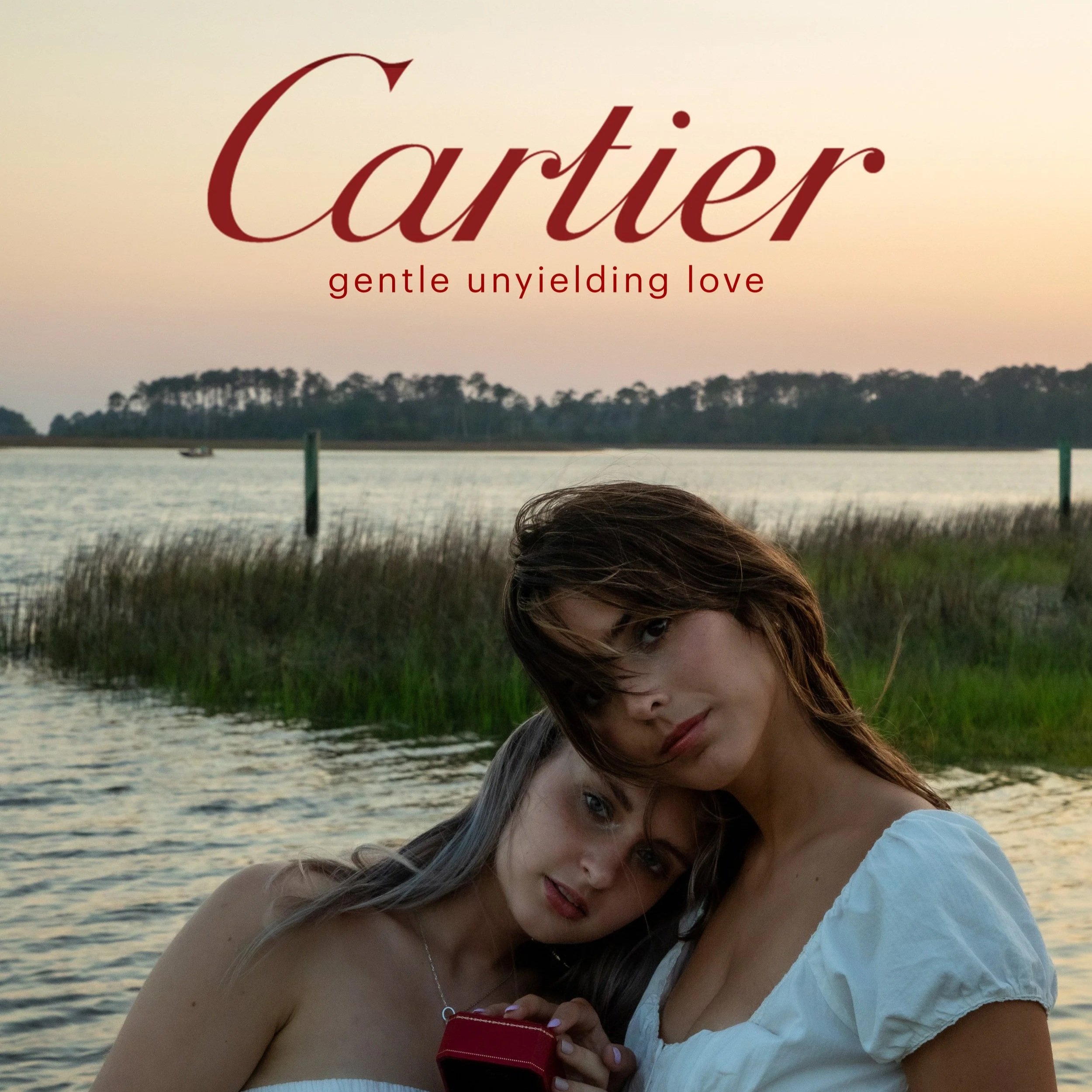 Two women with light skin and long brunette and blonde hair, close together by a body of water, holding a small red box, during sunset with trees in the background, with the text 'Cartier gentle unyielding love' at the top.