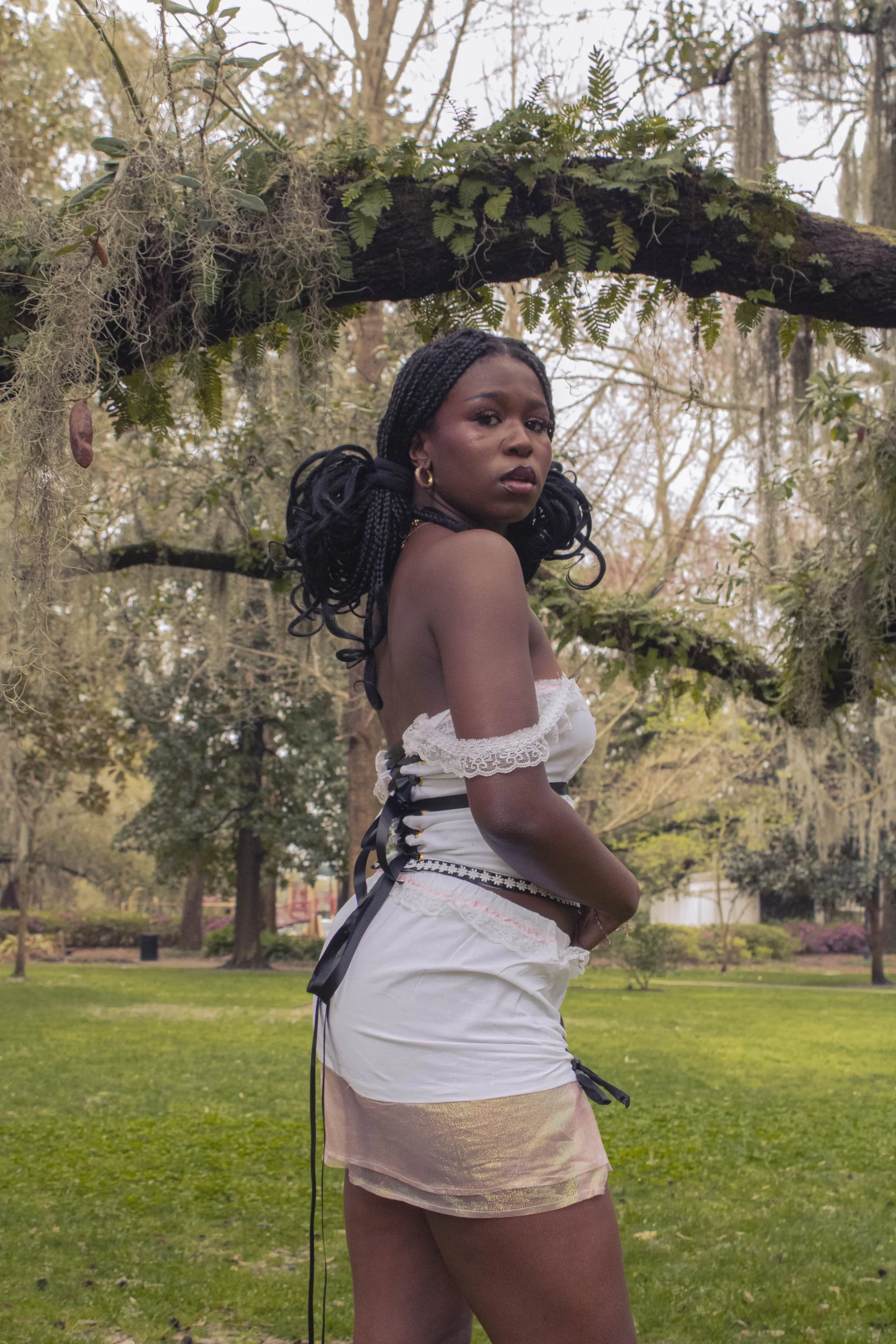 A woman with braided hair stands in a park, looking over her shoulder at the camera, wearing a white off-shoulder top and a white skirt with black accents. She's surrounded by trees and greenery, with some moss hanging from a branch above her.