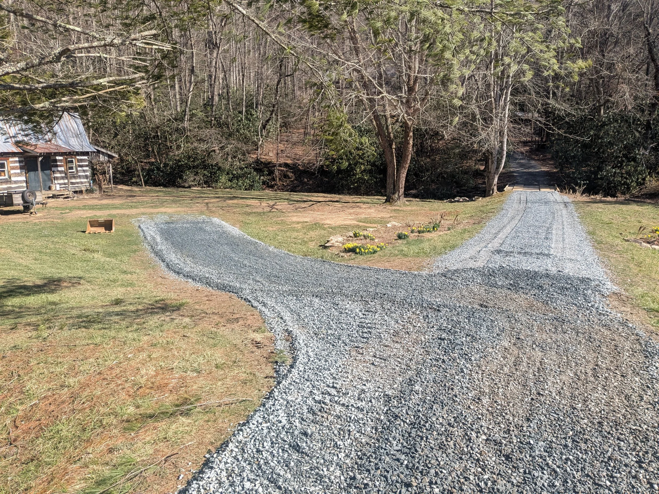 A gravel driveway forks into two paths in a rural yard, with an old wooden house to the left and trees in the background. There are small flower beds along the right side of the driveway.