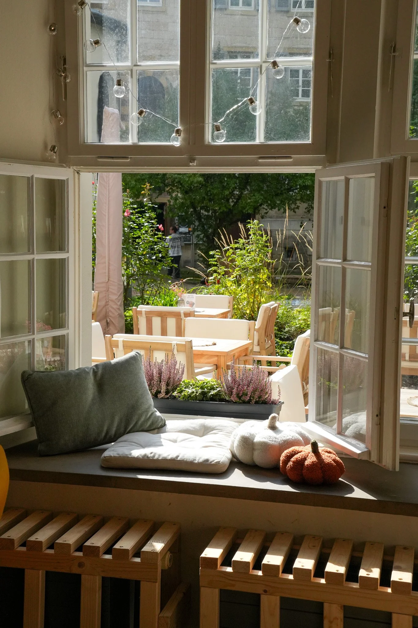 Blick aus einem offen stehenden Fenster auf einen Garten mit Holztischen und Gartenmöbeln, umgeben von Pflanzen und Bäumen.