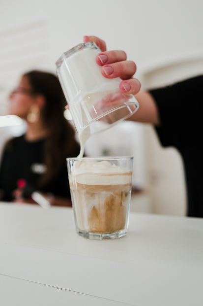 Person gießt Milch in einen Kaffee mit Schaum in ein Glas. Im Hintergrund eine Frau mit Brille.