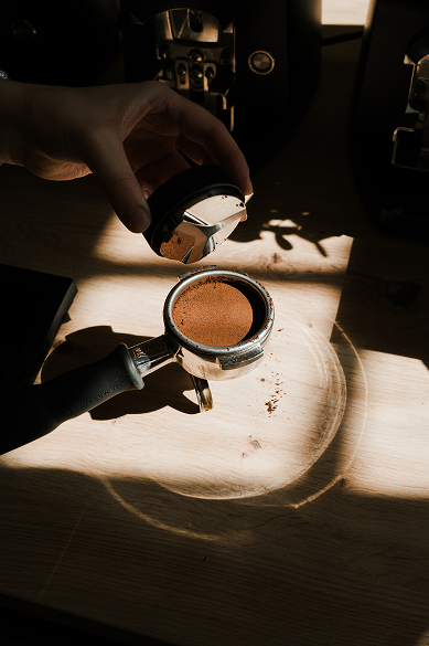 Hand, die Kaffeepulver in einen Espressomaschinen-Tamper füllt, auf einem Holzarbeitsplatz mit Espressomaschine im Hintergrund und Schatten.