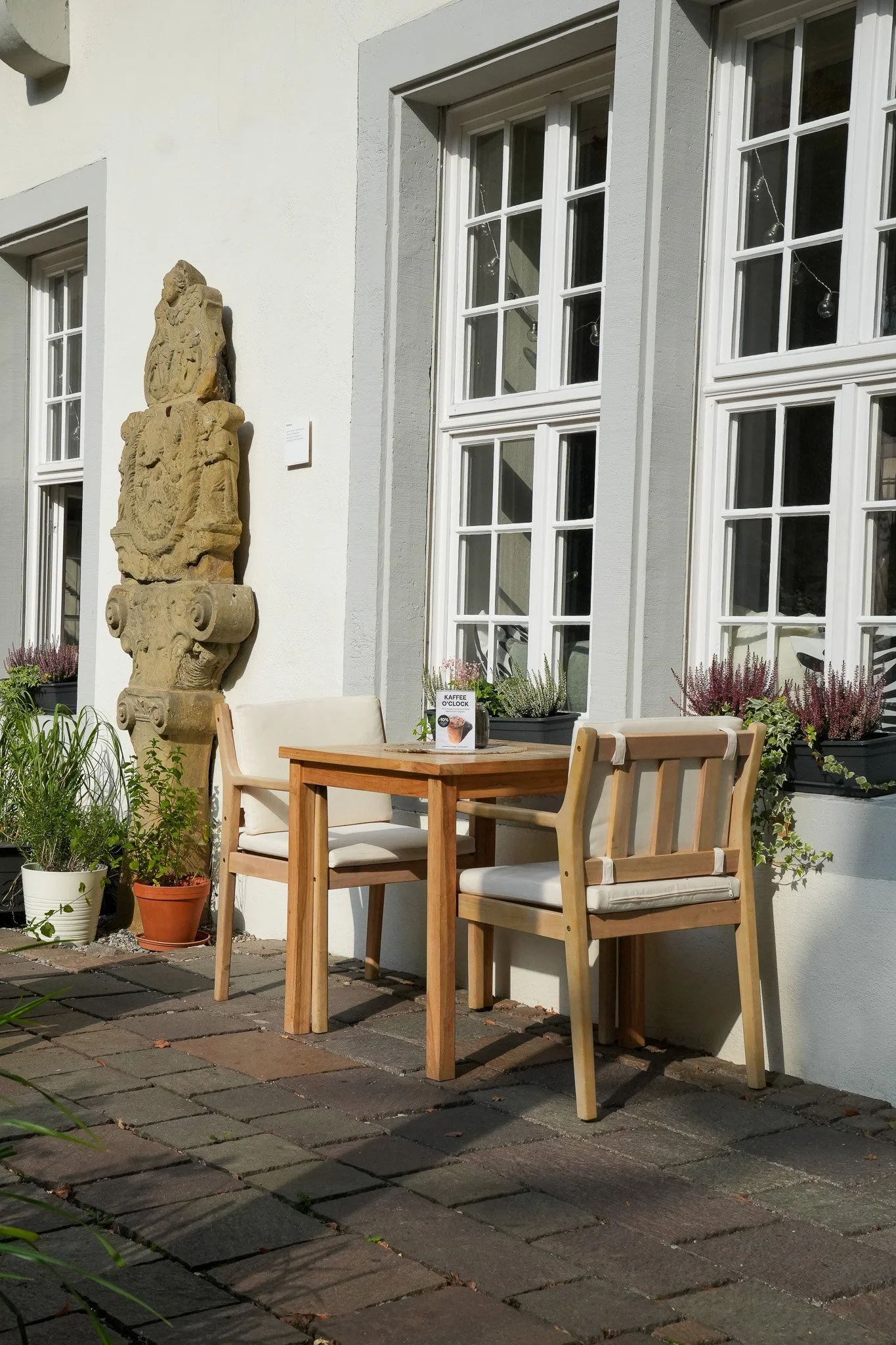 Ein kleiner Holztisch mit zwei Stühlen auf einer gepflasterten Terrasse vor einem weißen Haus mit großen Fenster. Es befinden sich Pflanzen in Töpfen und eine historische Steinskulptur an der Wand.