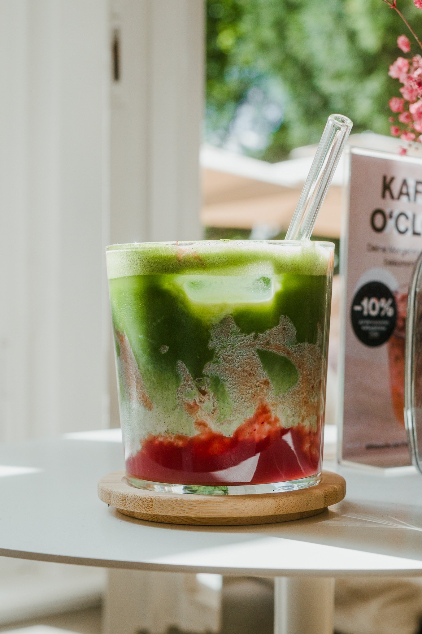 Ein Matcha Latte mit Erdbeerpüree, das auf einem kleinen hölzernen Untersetzer auf einem weißen Tisch steht. Im Hintergrund sind ein Fenster, ein Blumenstrauß und ein Werbeschild für Kaffee zu sehen.