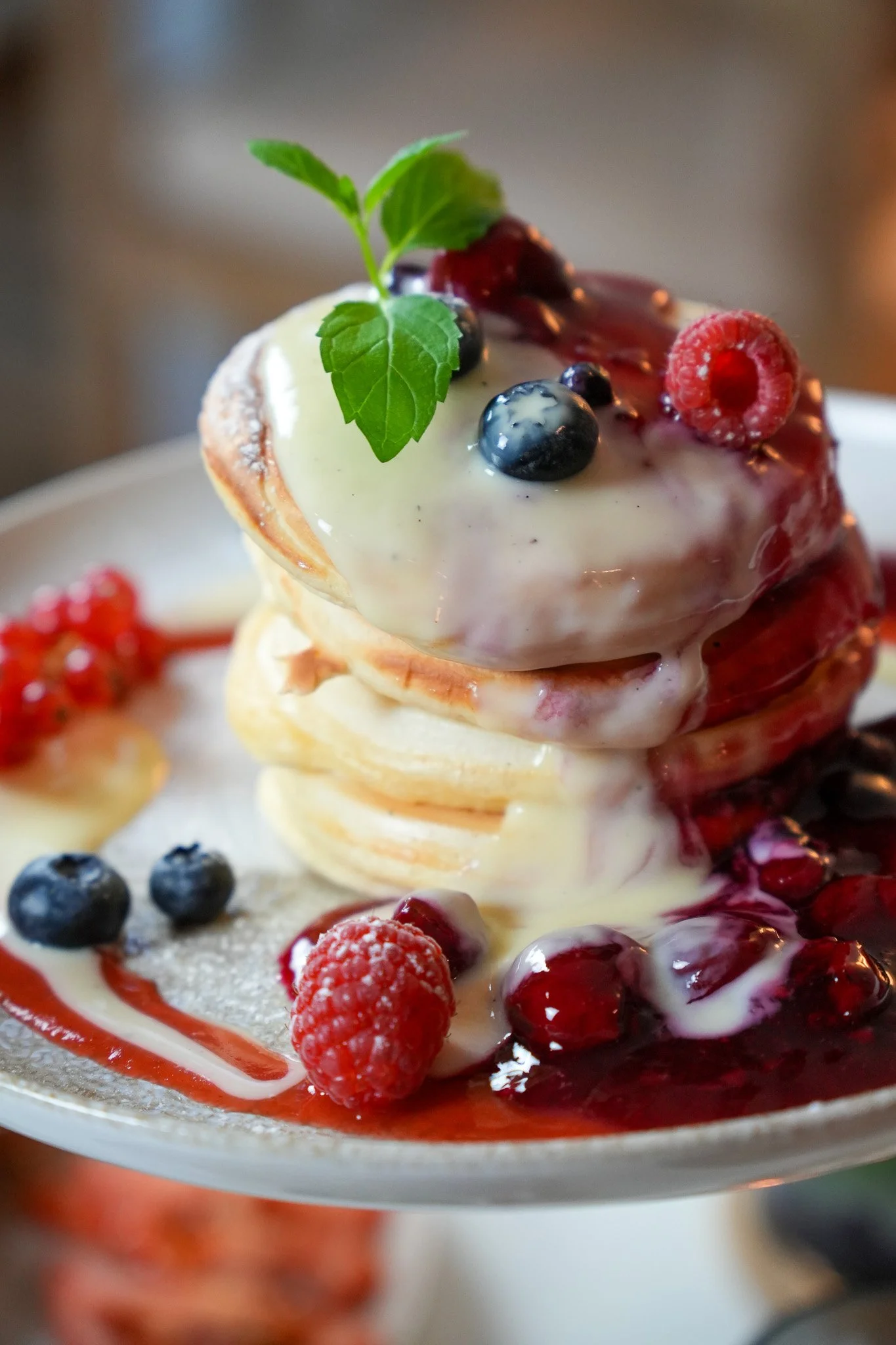 Ein Teller mit mehreren Pfannkuchen, garniert mit Himbeeren, Heidelbeeren, roten Johannisbeeren, einem Minzblatt und einer Vanillesoße, die über die Pfannkuchen läuft.