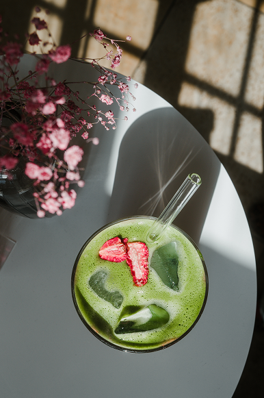 Ein grüner Smoothie mit Eiswürfeln und einer roten Blüte als Dekoration in einem Glas mit einem Strohhalm auf einem weißen Tisch, daneben eine Vase mit pinken Blumen, im Sonnenlicht.