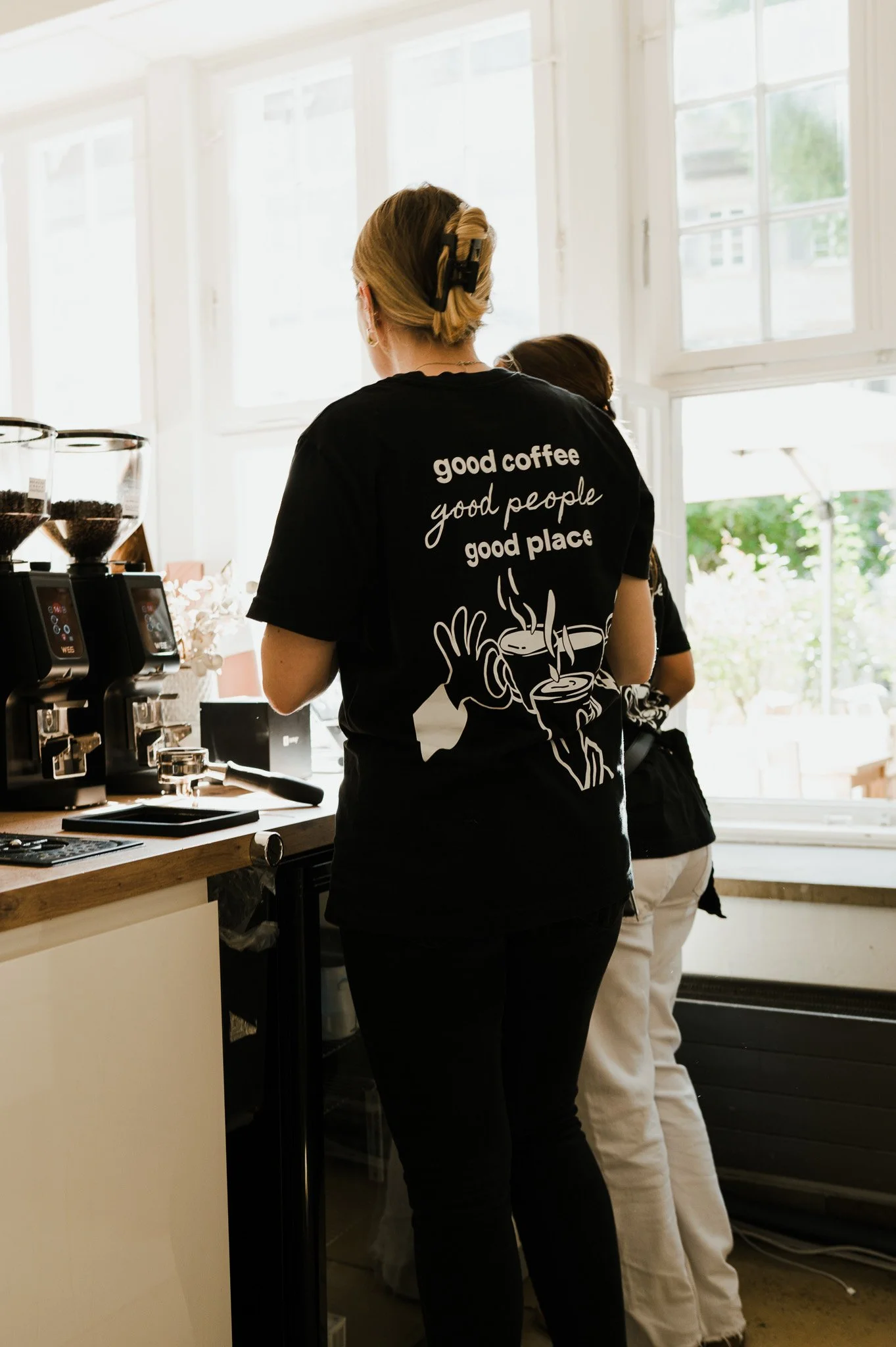 Zwei Frauen arbeiten in einer Kaffeebar, eine trägt ein schwarzes T-Shirt mit der Aufschrift 'good coffee, good people, good place' und einem Stehkaffee, die andere steht an einem Laib Selbstbedienungskaffeebereiter.