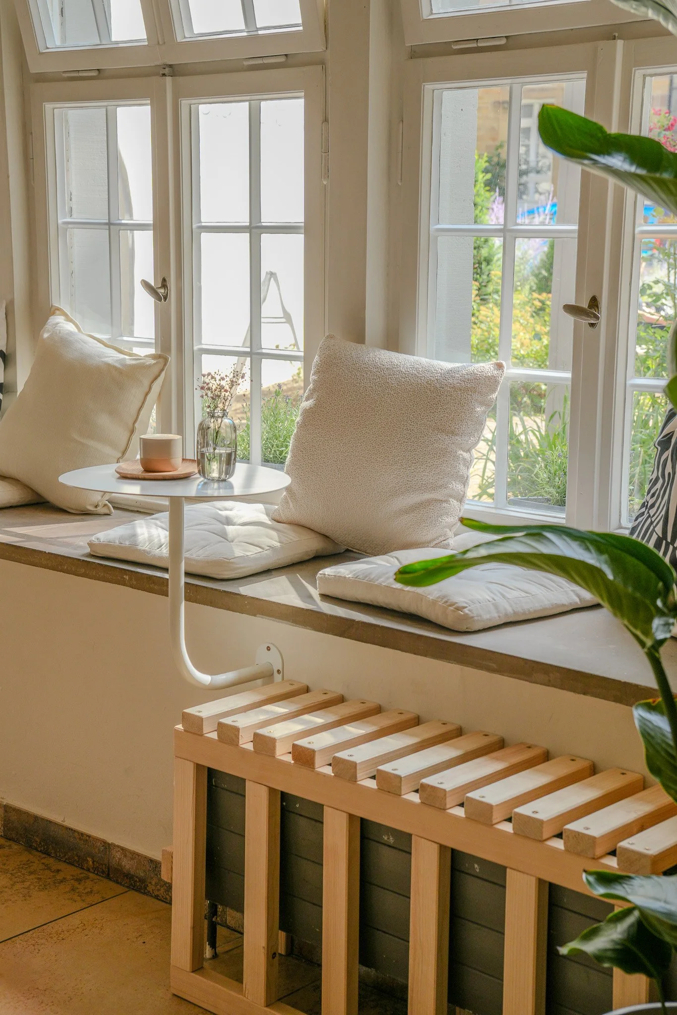 Gemütliche Sitzecke mit Kissen und einem kleinen Tisch am Fenster, mit Pflanzen und natürlichem Licht, darunter ein Heizkörper mit Holzabdeckung.