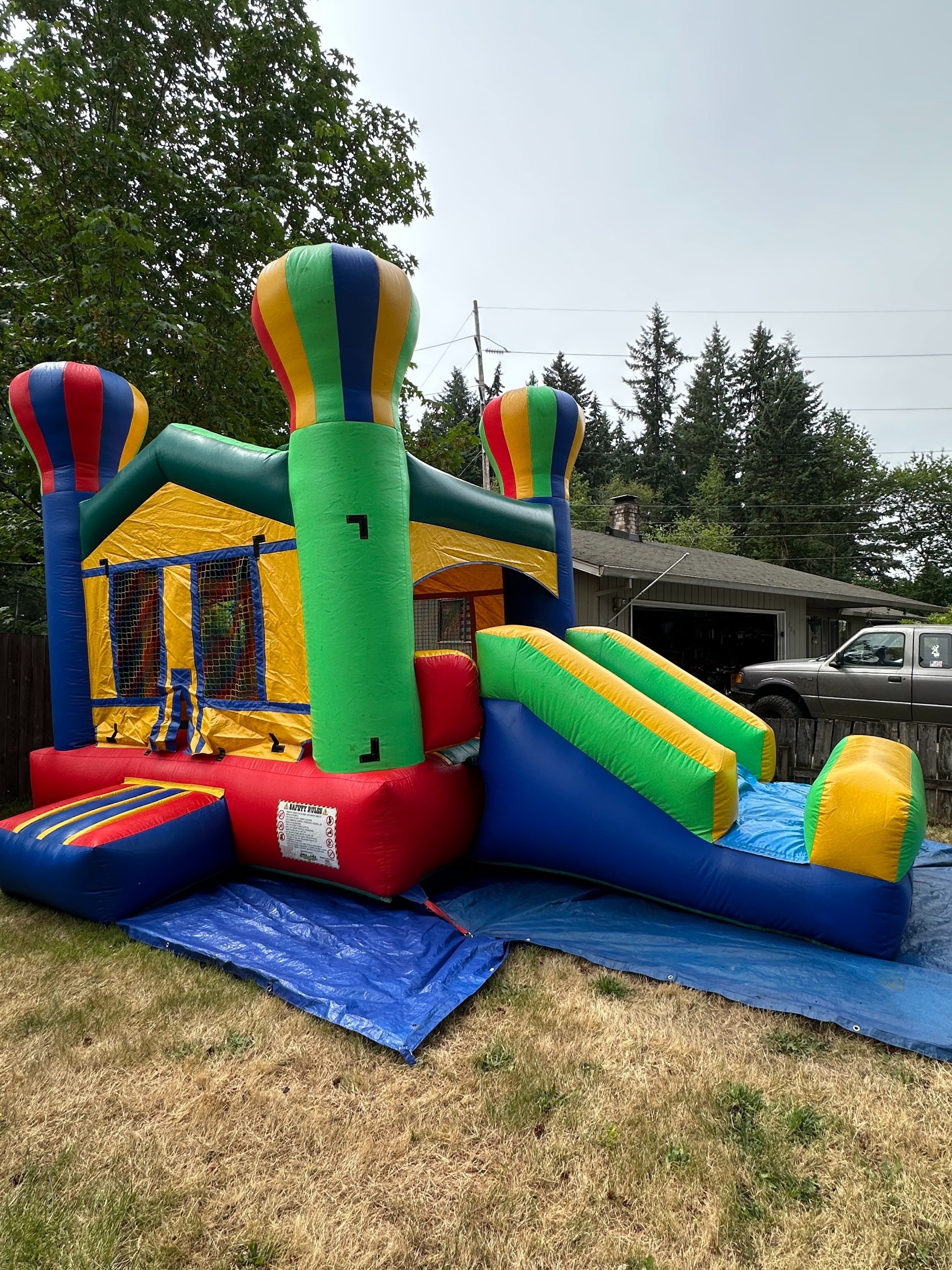 Colorful inflatable bounce house with slide outside on a grassy yard, surrounded by trees and a house in the background.