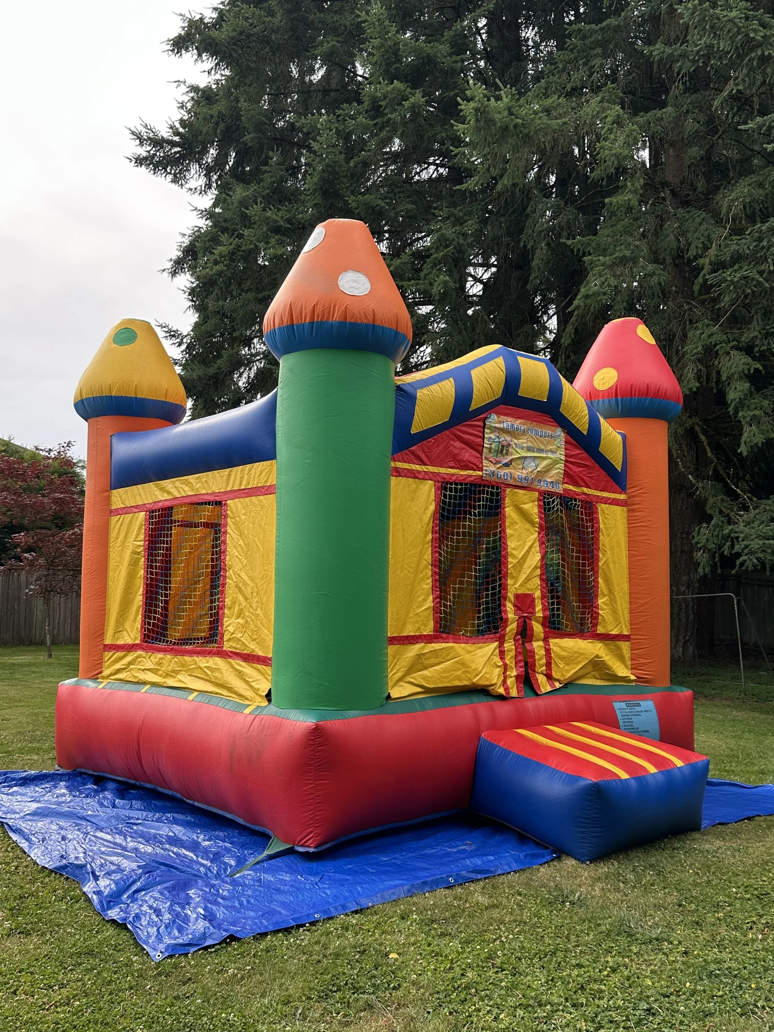 Colorful inflatable bounce house designed to resemble a castle with turrets, set up on a blue tarp in a grassy backyard with evergreen trees in the background.