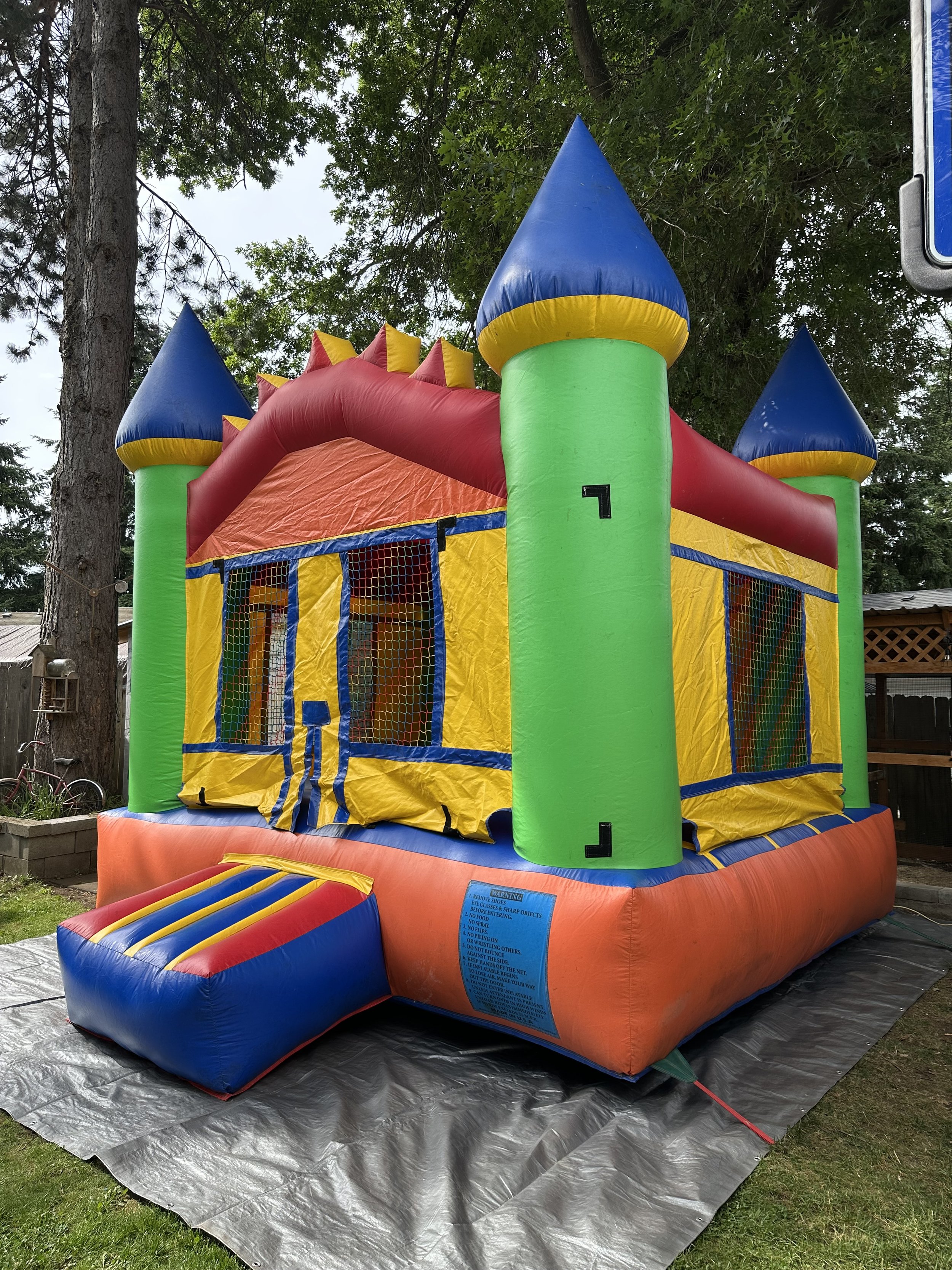 Colorful inflatable bounce house with four blue-topped towers, yellow and red walls, a small ramp at the entrance, and netted windows, set outdoors on a tarp.