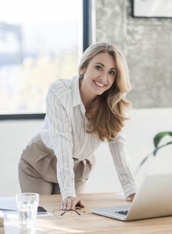 A smiling woman with long blonde hair, wearing a white striped blouse, leaning over a desk in an office with large windows and a brick wall in the background.