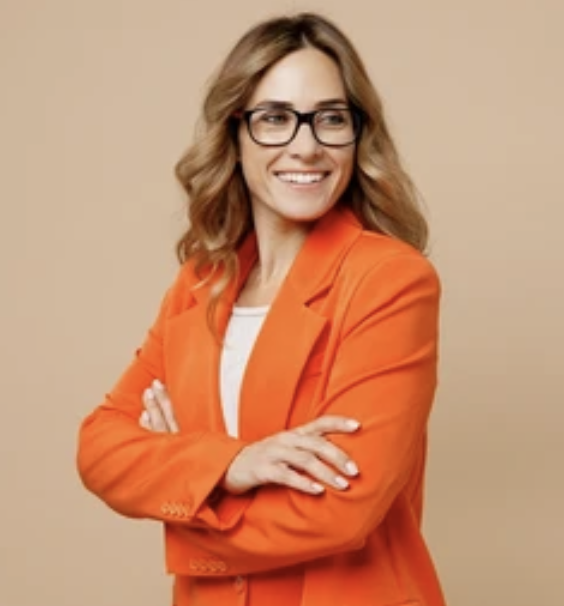A woman with wavy blonde hair, wearing glasses and an orange blazer, smiling with arms crossed against a beige background.
