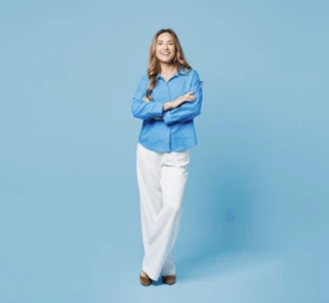 A smiling woman stands against a blue background, crossed arms, wearing a blue jacket and white pants.