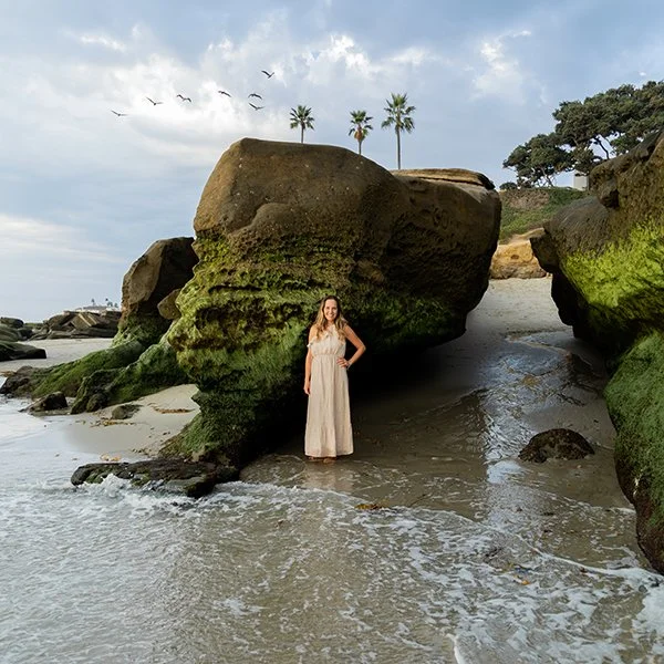 Woman in a long beige dress standing on a sandy beach with large moss-covered rocks and palm trees in the background, under a partly cloudy sky with birds flying.