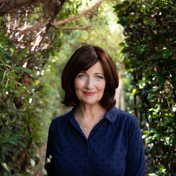 A woman with shoulder-length dark brown hair and blue eyes, wearing a navy blue blouse, standing outdoors among green trees and foliage.