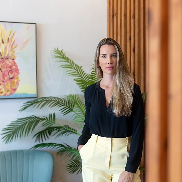 A woman with long blonde hair, wearing a black blouse and yellow pants, standing indoors next to a green plant and a colorful pineapple painting on the wall.
