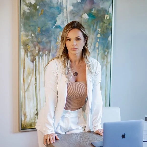 A woman with wavy blonde hair, wearing a white blazer and beige top, stands behind a wooden table with a closed silver MacBook laptop, in front of an abstract colorful painting in an office setting.