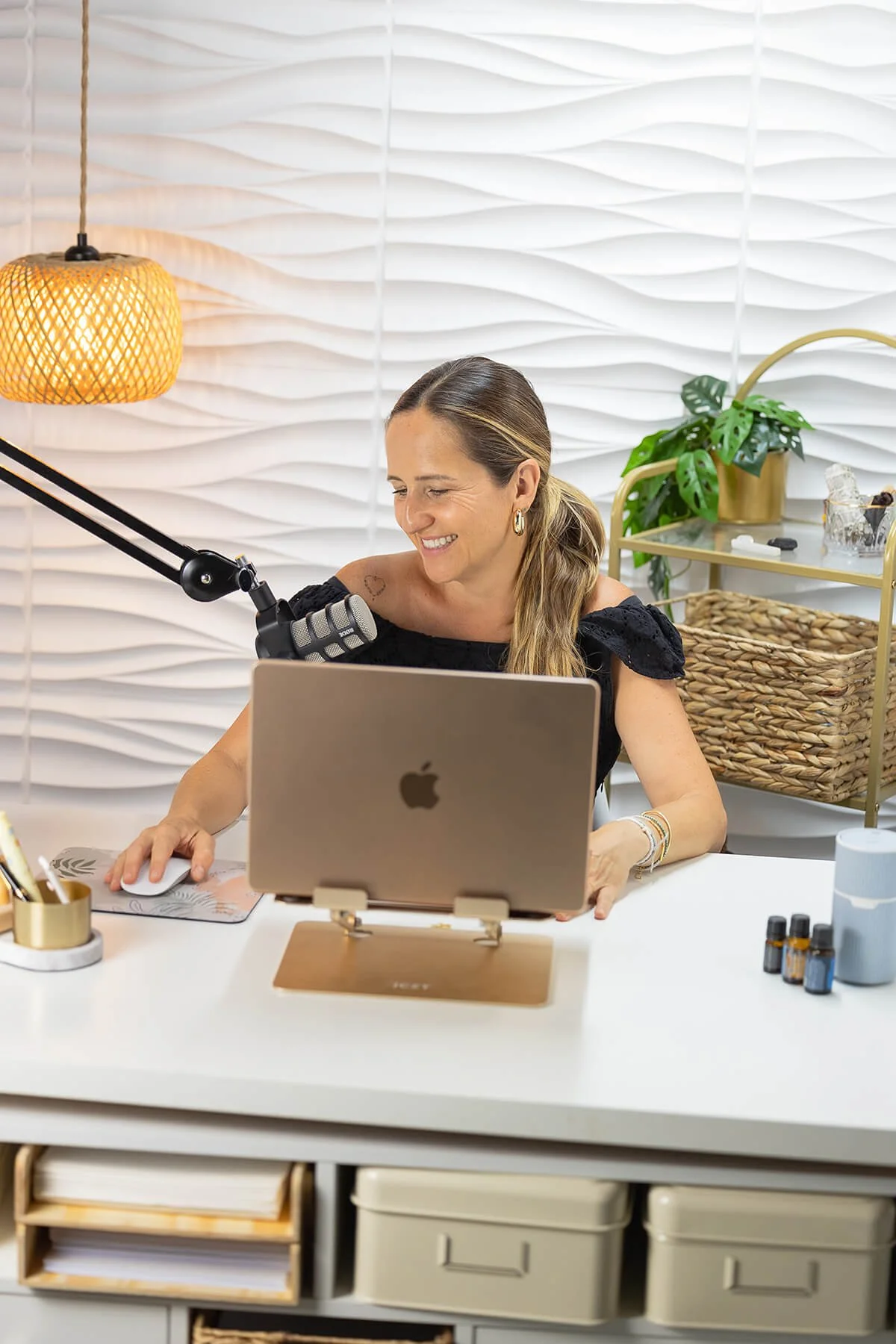 A woman with long hair wearing a black top, sitting at a white desk, smiling while working on a laptop with a microphone attached, in a modern room with white textured wall, hanging light, plant, and office supplies.