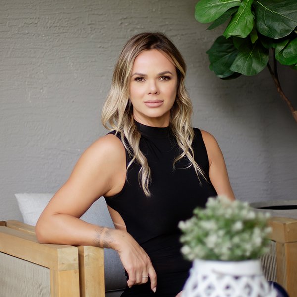 A woman with blonde hair wearing a sleeveless black top, sitting on a wooden chair indoors, with a green plant and a white pot in the foreground.