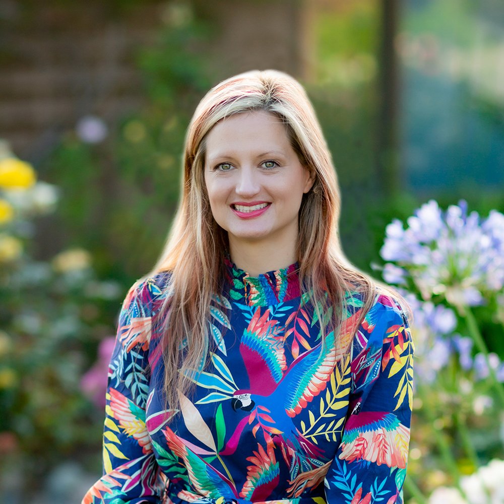 A woman with blonde hair smiling outdoors in a garden, wearing a colorful, patterned dress with birds and foliage.