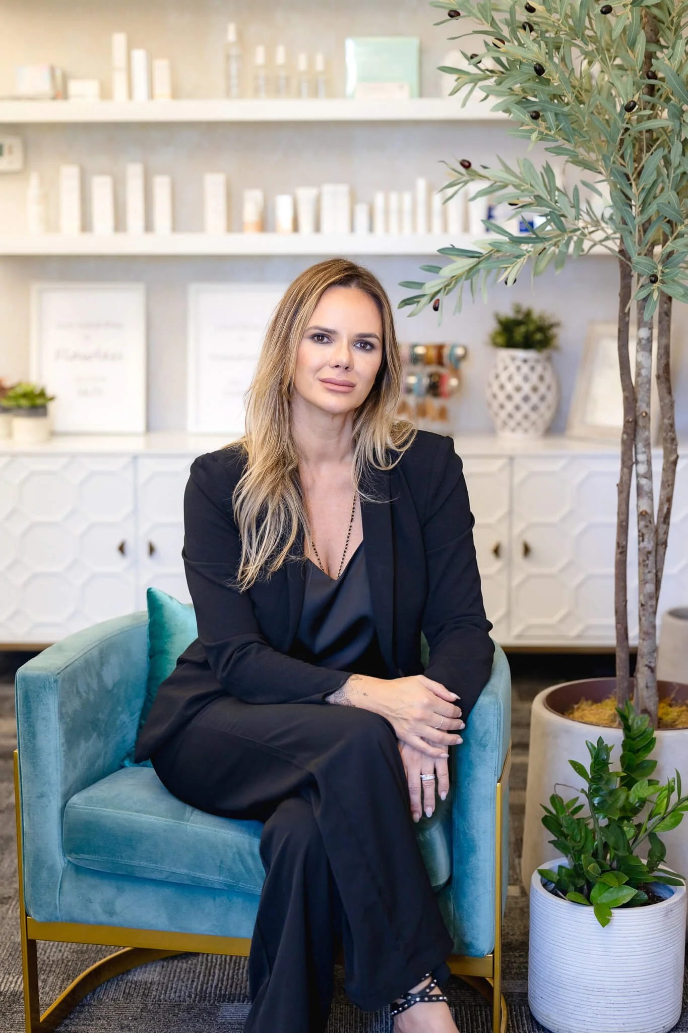 Woman in black blazer sitting on teal armchair in modern interior with shelves, plants, and framed artwork