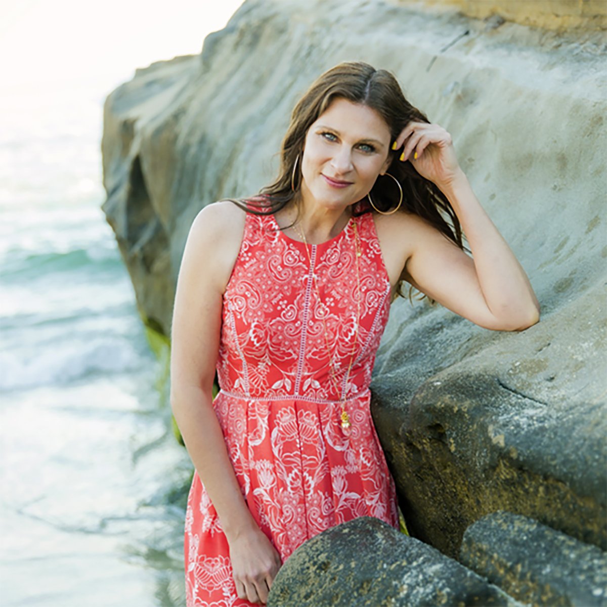 A woman with long brown hair, wearing a bright coral dress with white floral patterns, leaning against a large rock by the ocean on a sunny day.