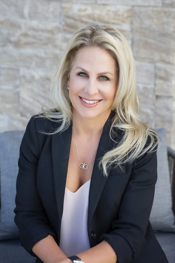 A blonde woman in a black blazer and white top, smiling while sitting against a brick wall background.