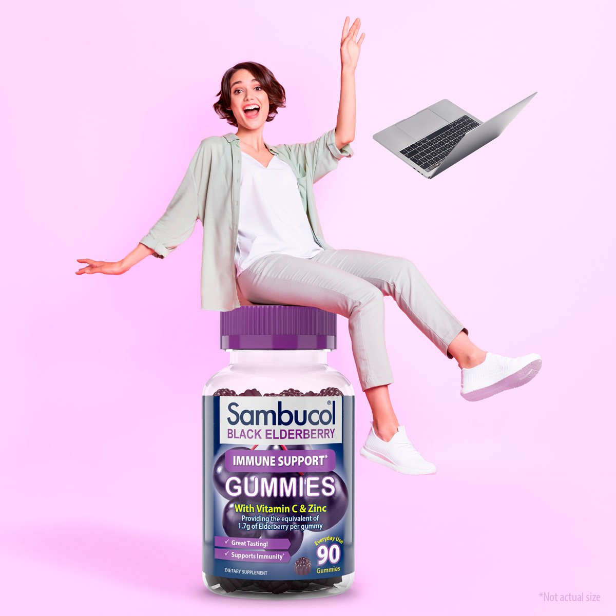 A woman sitting on a large Sambucol black elderberry gummies bottle, with a laptop floating nearby, against a pink background.